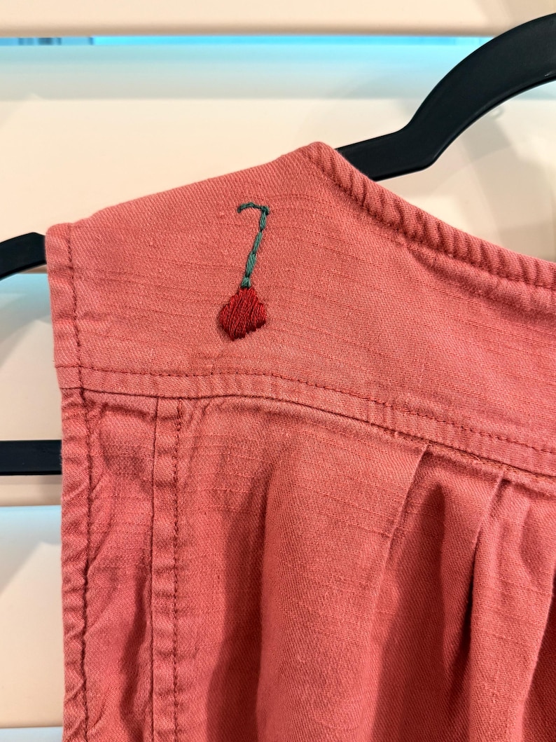 Puede incluir: Primer plano de una prenda de color rosa empolvado con un peque&ntilde;o dise&ntilde;o floral bordado en rojo y verde en el hombro. La tela tiene una textura visible y est&aacute; colgada de una percha negra.