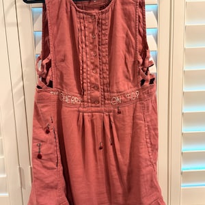 Puede incluir: Vestido sin mangas, color rosa empolvado, con botones en la parte delantera y pliegues decorativos. El vestido presenta cortes laterales y texto bordado que dice "THE CHERRY ON TOP". Peque&ntilde;os adornos en forma de cereza est&aacute;n cosidos en la parte delantera.
