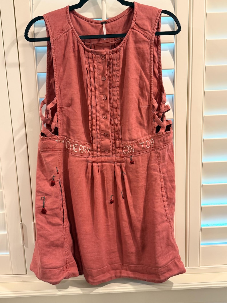 Puede incluir: Vestido sin mangas, color rosa empolvado, con botones en la parte delantera y pliegues decorativos. El vestido presenta cortes laterales y texto bordado que dice "THE CHERRY ON TOP". Peque&ntilde;os adornos en forma de cereza est&aacute;n cosidos en la parte delantera.