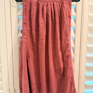 Puede incluir: Vestido sin mangas, color rosa empolvado, colgado de una percha negra. El vestido presenta un corpi&ntilde;o plisado, bolsillos laterales y costuras decorativas. Un peque&ntilde;o detalle bordado en rojo es visible cerca del hombro.