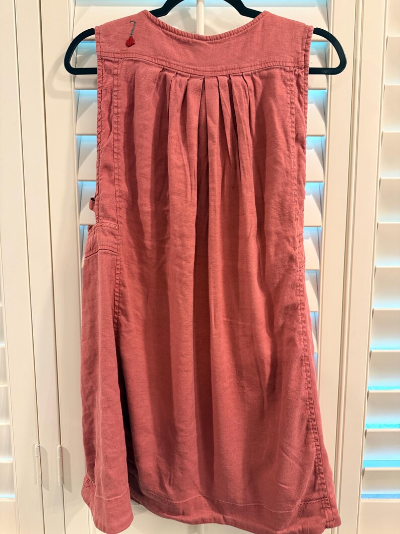 Puede incluir: Vestido sin mangas, color rosa empolvado, colgado de una percha negra. El vestido presenta un corpi&ntilde;o plisado, bolsillos laterales y costuras decorativas. Un peque&ntilde;o detalle bordado en rojo es visible cerca del hombro.
