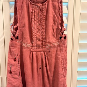 Puede incluir: Vestido delantal rosa polvoriento con botones en la parte delantera y cortes laterales. El texto "THE CHERRY ON TOP" est&aacute; bordado en la parte delantera. Peque&ntilde;os detalles de cerezas bordados en la parte delantera y los lados.