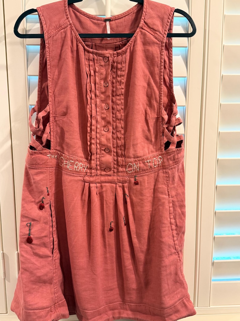 Puede incluir: Vestido delantal rosa polvoriento con botones en la parte delantera y cortes laterales. El texto "THE CHERRY ON TOP" est&aacute; bordado en la parte delantera. Peque&ntilde;os detalles de cerezas bordados en la parte delantera y los lados.
