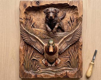 Könnte beinhalten: Ein geschnitztes Holzrelief mit einem Labrador Retriever und einer Ente im Flug. Der Hund befindet sich über der Ente, mit Schilf und Wasserelementen. Der Text "CNC & 3D PRINT READY" und "Dog Duck Hunting STL 3D Relief STL" sind sichtbar.