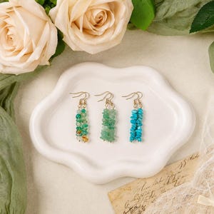 May include: Three pairs of dangle earrings featuring small, irregular, tumbled gemstone chips in shades of green and blue. Each earring is suspended from a gold-toned metal hook. The stones vary in color from light turquoise to deep emerald green.