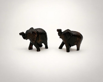 May include: Two small, hand-carved wooden elephant figurines with a dark brown, polished finish. Each elephant is depicted in a standing position with its trunk slightly raised, showcasing visible wood grain textures and carved details.