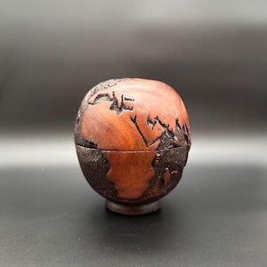 May include: A wooden globe-shaped box with a detailed carving of the world map. The box is made of reddish-brown wood and has a textured, dark brown base. The top of the box opens to reveal a hollow interior.