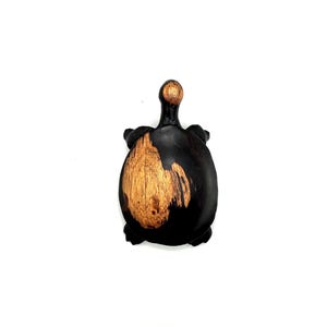 May include: A small turtle figurine carved from wood, featuring a smooth, dark brown finish across the body and limbs, with a prominent, light tan, natural wood grain patch on the centre of the shell and a matching light tan accent on the head.
