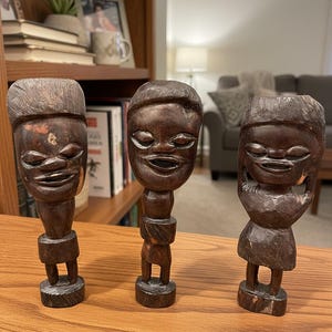 May include: Three dark brown wooden sculptures of stylized human figures with smiling faces. Each figure has a unique carved design, including headwear and body details. The sculptures are displayed on a wooden surface.
