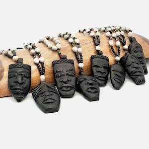 May include: A collection of seven necklaces featuring dark brown carved wooden pendants shaped like human faces. Each pendant is suspended from a cord strung with white, black, and small red and green beads, creating a uniform design across the set.