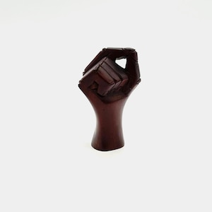 May include: A carved wooden sculpture of a fist, with the hand clenched in a fist. The sculpture is a dark brown color and has a smooth finish. The fist is raised in a gesture of solidarity.