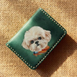 Könnte beinhalten: Grünes Lederportemonnaie mit einem detaillierten Porträt eines flauschigen, hellfarbigen Hundes mit dunklen Augen und einem bunten gestreiften Kragen. Das Portemonnaie hat weiße Nähte an den Rändern und einen strukturierten, beigefarbenen Hintergrund.