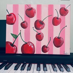 Puede incluir: Pintura sobre lienzo con cerezas rojas y tallos verdes sobre un fondo a rayas rosas y blancas. La firma "Katy L" es visible. La obra está colocada sobre un piano negro. El estilo de la pintura recuerda al pop art.