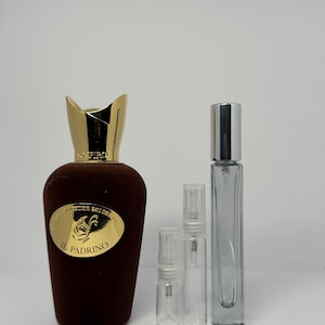 Puede incluir: Un frasco de perfume marrón con tapón y etiqueta dorados, junto a tres pequeños frascos pulverizadores transparentes y un frasco rectangular alto. La etiqueta dice "IL Padrino".