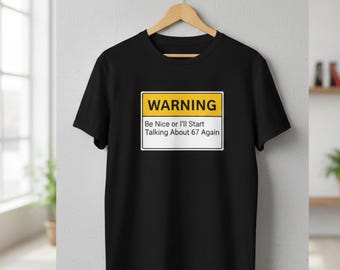 Può includere: T-shirt nera con una grafica rettangolare gialla e bianca con la scritta "WARNING" e il testo "Be Nice or I'll Start Talking About 67 Again". La maglietta è appesa a una gruccia di legno.