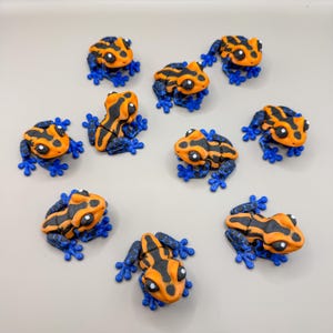 Può includere: Una collezione di piccole figurine decorative a forma di rana. Ogni rana presenta un corpo arancione vibrante con strisce nere e zampe blu. Le rane sono disposte su una superficie grigio chiaro, mostrando il loro design dettagliato e i colori vivaci.