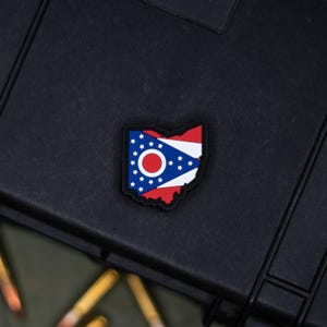 May include: A colorful patch in the shape of Ohio, featuring the state flag design. The patch has a red border, a white and blue section with white stars, and a red and white circle in the center. The patch is on a black surface.
