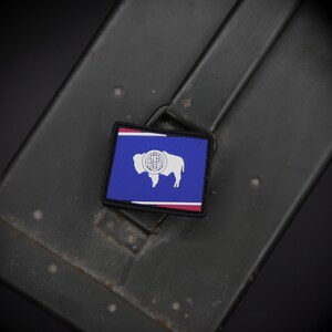 May include: A square patch featuring the Wyoming state flag, with a blue background, white bison, and red and white borders. The patch is set against a dark, textured background.