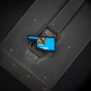 May include: A patch featuring the outline of the state of Oklahoma in blue, with the state seal in the center. The word "OKLAHOMA" is printed in white. The patch is set against a dark, textured background.