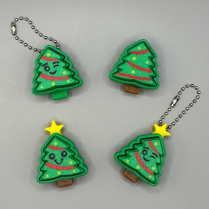 May include: Four green Christmas tree keyrings with red and yellow detailing. Each tree has a brown trunk and a silver chain. Two trees have winking faces, and two have a yellow star on top.