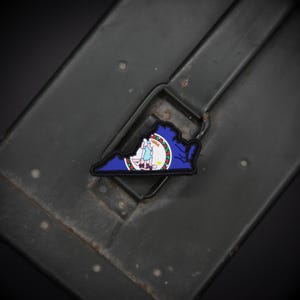 Könnte beinhalten: Ein Aufnäher in Form des Bundesstaates Virginia, mit blauem Rand und einem detaillierten Emblem mit einer Frau, einem Speer und einem Banner. Der Aufnäher befindet sich auf einem dunklen, strukturierten Hintergrund.