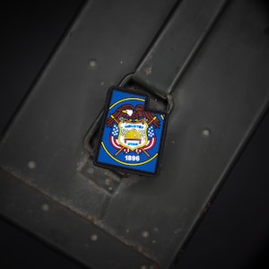 Puede incluir: Un parche rectangular con el emblema del estado de Utah. El parche tiene un borde azul y un diseño central con un escudo, un águila y la palabra "UTAH". El año "1896" se muestra en la parte inferior. El parche está sobre un fondo oscuro y texturizado.