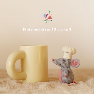 May include: A cream-coloured mug with a rounded handle sits beside a crocheted mouse wearing a chef's hat. The mouse is grey with pink accents and is approximately 10 cm tall. The text reads "Finished size: 10 cm tall".