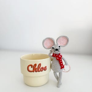 May include: A cream-colored mug with the name "Chloe" in red script is next to a crocheted gray mouse wearing a red scarf. The mouse has pink inner ears and a pink tail. The mug is on a white surface.