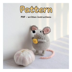 Könnte beinhalten: Eine gehäkelte graue Maus, die ein gelbes Käse-Stück hält, mit rosa Ohren und einem rosa Schwanz. Neben der Maus liegt eine Knoblauchzehe. Der Text "Pattern" und "PDF - written instructions" sind oben.