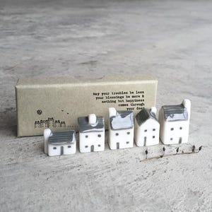 Peut inclure: Une collection de maisons miniatures en céramique avec des toits gris et des murs blancs. Les maisons sont disposées en rangée, avec une petite boîte en carton derrière elles. La boîte porte l'inscription "May your troubles be less..."