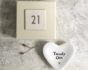 May include: A cream-colored gift box with the number "21" on a square tile, and a white heart-shaped dish with the words "Twenty One" and a small heart symbol. A sprig of dried plant material is also present.