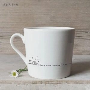 Könnte beinhalten: Weißer Keramikbecher mit gebogenem Henkel. Der Becher zeigt eine schwarze Illustration eines Dorfes und den Text "Tea is a warm little hug in a mug..." Der Becher misst 8 cm x 7,5 cm.