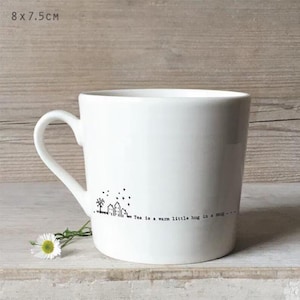 Puede incluir: Taza de cerámica blanca con asa curva. La taza presenta una ilustración negra de un pequeño pueblo con un árbol y estrellas, y el texto "Tea is a warm little hug in a mug...". La taza mide 8 cm x 7,5 cm.