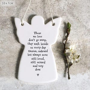 Könnte beinhalten: Weißes Keramik-Engelorna-ment mit dem Text: "Die, die wir lieben, gehen nicht weg, sie gehen jeden Tag an unserer Seite." Das Ornament ist 10 cm x 7 cm groß. Ein Zweig mit weißen Blüten liegt neben dem Engel.