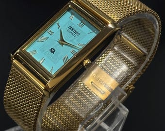 May include: A gold-toned Seiko Quartz watch with a rectangular turquoise face and Roman numeral hour markers. The watch features a mesh band and is displayed on a clear stand. The watch face has the Seiko Quartz logo.
