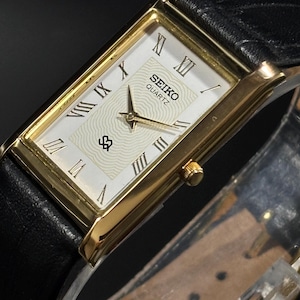 Puede incluir: Reloj Seiko Quartz rectangular dorado con esfera blanca y correa de cuero negro. La esfera del reloj presenta números romanos y agujas doradas. Diseño clásico.