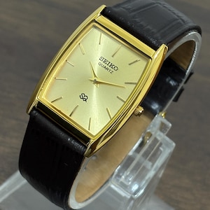 Puede incluir: Reloj Seiko Quartz dorado con esfera rectangular y correa de cuero negro. La esfera del reloj es de color oro claro con agujas y marcadores negros. El reloj se muestra sobre un soporte transparente.
