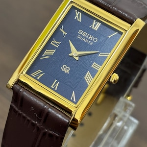 May include: A rectangular Seiko Quartz watch with a gold-tone case and a dark blue dial. The dial features gold Roman numerals and hands. The watch has a brown leather strap with a crocodile pattern.