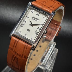 May include: A rectangular Seiko Quartz watch with a silver case and a light-coloured face with Roman numerals. It has a brown leather strap with a crocodile pattern. The watch is displayed on a clear stand against a dark background.