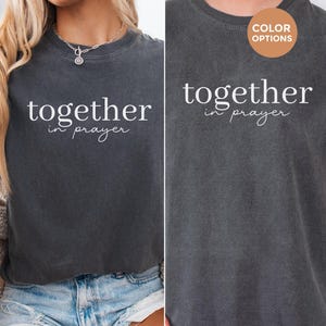 May include: Two charcoal gray t-shirts with the text "together in prayer" in white. One shirt is worn by a person wearing denim shorts and a necklace. The other shirt is worn by a person with a brown circle that says "color options".