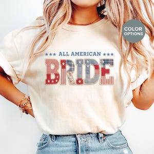 May include: A cream-colored short-sleeve t-shirt featuring the text ALL AMERICAN BRIDE in blue. The word BRIDE is designed with a patchwork pattern in red, white, and blue, incorporating stars, stripes, and floral motifs with visible stitching.