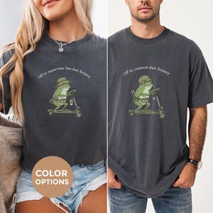 May include: Two dark gray t-shirts with a frog graphic riding a scooter. The frog wears a hat and has a coffee cup. Text above the frog reads "Off to supervise the dad foolery" and "Off to commit dad foolery". A color options graphic is in the image.
