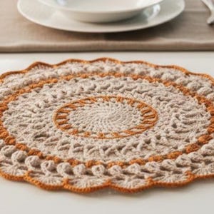 May include: A handmade, beige crochet placemat with an orange border, set on a white table. The circular mat is surrounded by silverware and a white plate and bowl. The placemat is approximately 14 inches in diameter.