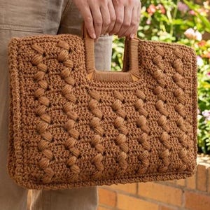 May include: A brown, hand-crocheted tote bag with a textured pattern and wooden handles. The bag is rectangular with a structured design, perfect for carrying essentials. The bag is held by a person in a natural outdoor setting.