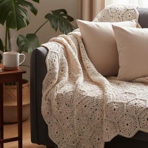 May include: A cream-coloured, crocheted blanket with a scalloped edge is draped over a dark grey sofa. Two beige square cushions rest against the back of the sofa. A mug sits on a small wooden table.