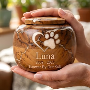 May include: A wooden pet urn with a paw print and heart design. The urn is brown with a light-colored grain pattern and a wooden lid. The name "Luna" and dates 2008-2023 are engraved on the front, along with the phrase "Forever By Our Side."