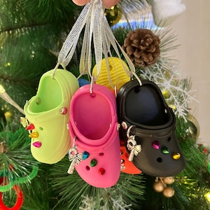 Puede incluir: Un conjunto de adornos navideños en miniatura con forma de zapatos, colgados de cintas plateadas. Los adornos, en colores vibrantes como verde, rosa, amarillo, negro y naranja, están decorados con abalorios y gemas. Se exhiben frente a un árbol de Navidad.