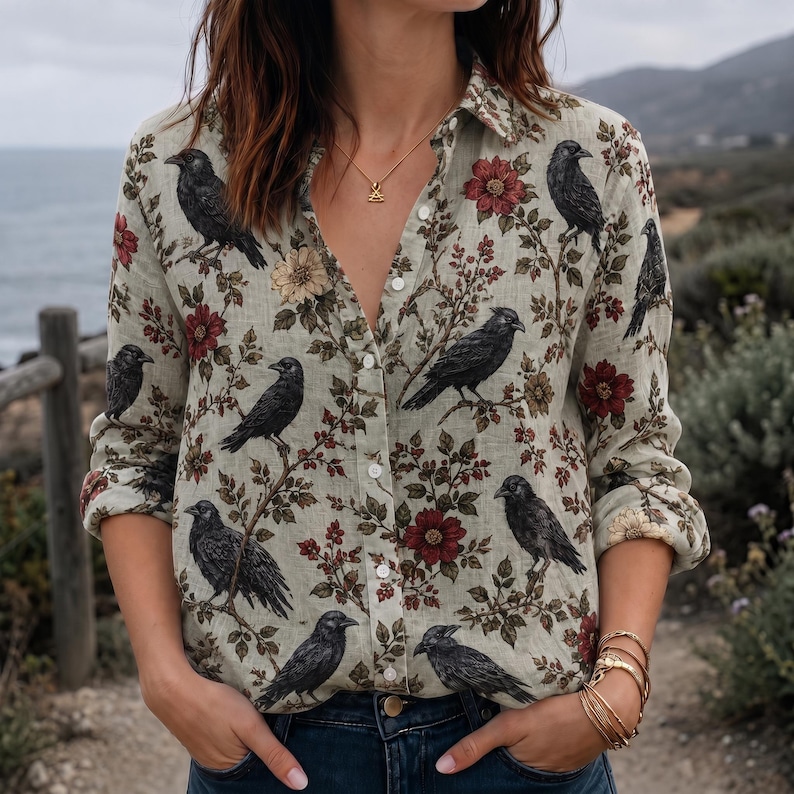 May include: A long-sleeve button-down shirt in a light beige color featuring a repeating pattern of black crows perched on branches with dark red and muted yellow flowers. The shirt has a classic collar and white buttons down the front placket.