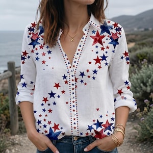 May include: A white long-sleeve button-down shirt featuring a scattered pattern of red and blue stars. The shirt has a classic collar, a front button placket, and long sleeves with cuffs. The star print varies in size across the fabric.