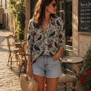 May include: A long-sleeved button-down shirt featuring a repeating pattern of black birds and red flowers on a light gray background, paired with light blue denim shorts with a raw hem and a woven straw handbag with a circular handle.